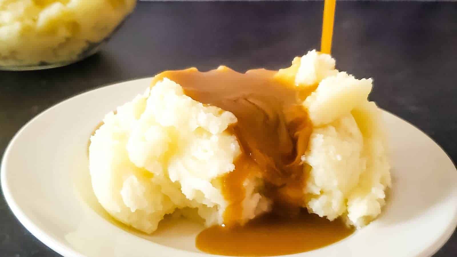 A plate of creamy mashed potatoes topped with rich brown gravy on a dark table. In the background, there is a glass bowl containing more mashed potatoes.
