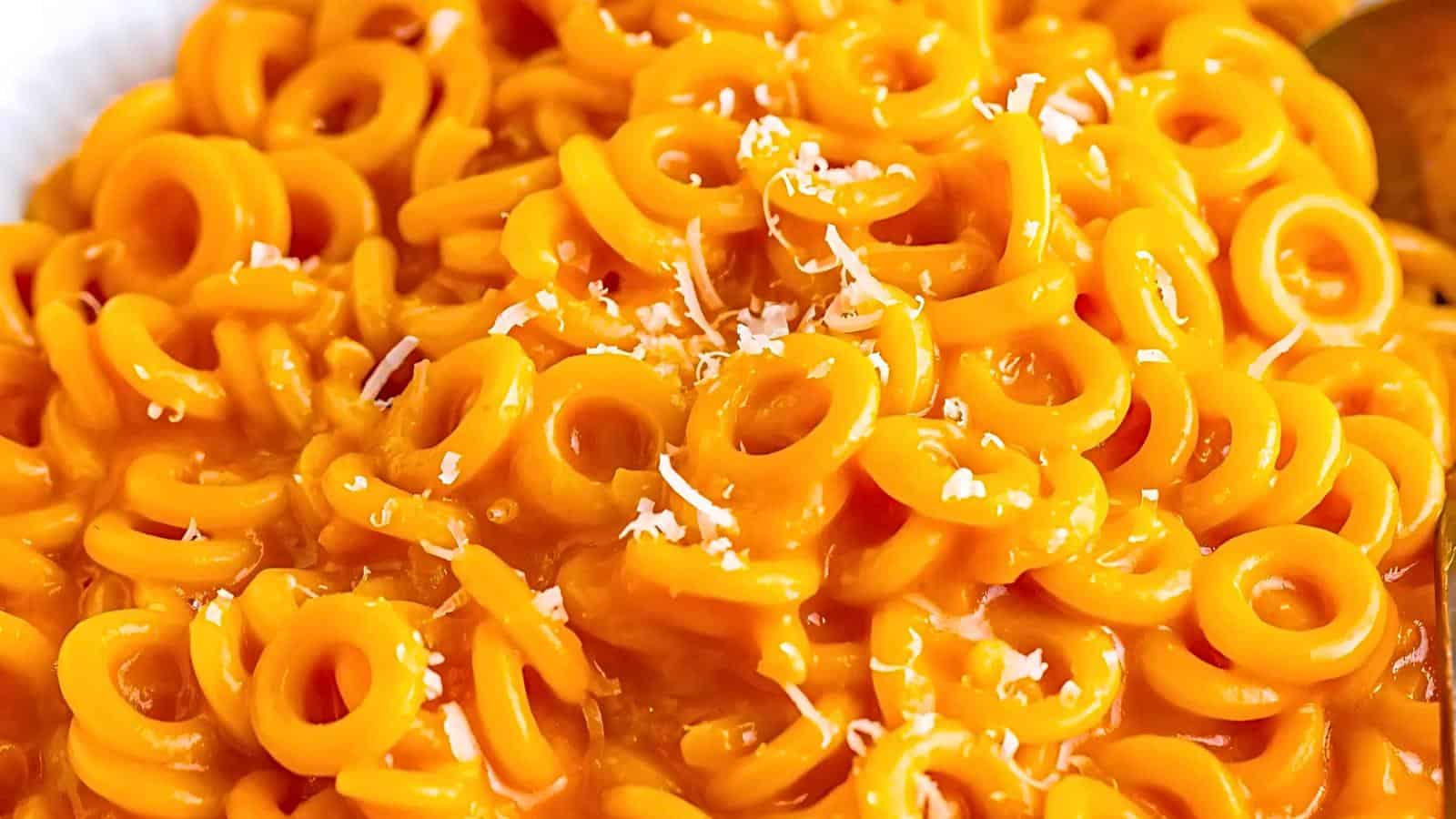 Close-up of a plate filled with creamy ring-shaped pasta, coated in a rich, vibrant orange cheese sauce and garnished with a light sprinkle of shredded cheese.