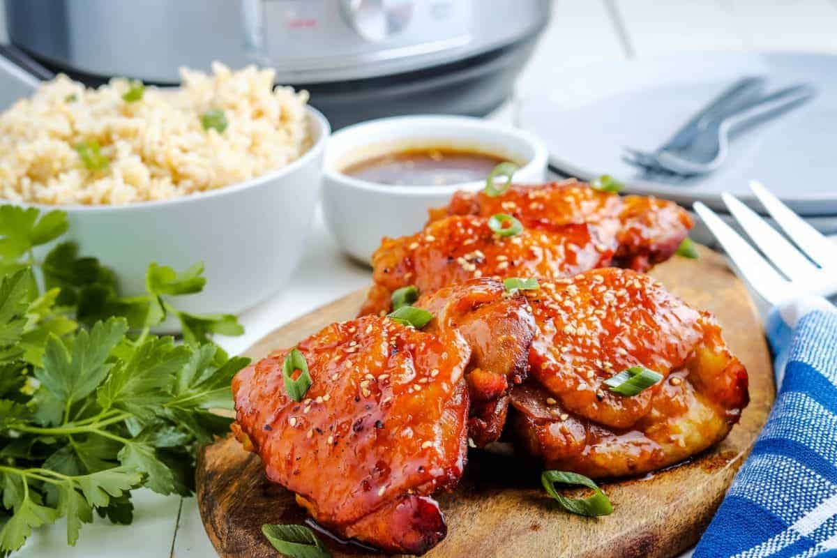 A plate featuring glazed chicken thighs garnished with chopped green onions sits on a wooden board. A white bowl of steamed rice is in the background, next to a cup of brown sauce and a small bunch of fresh parsley. A fork and knife lie beside a blue napkin.