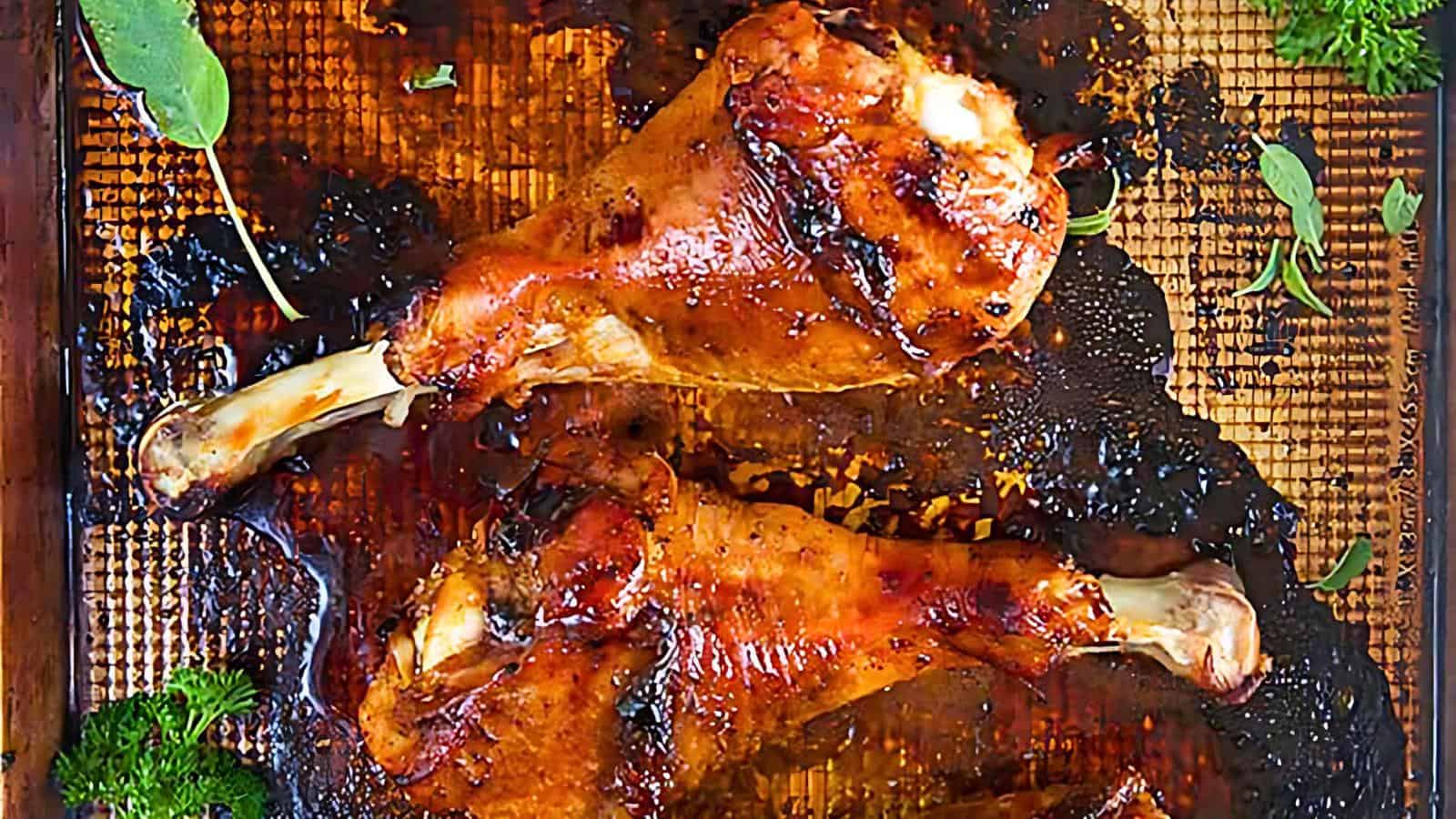 A close-up of roasted turkey legs placed on a baking sheet. The legs are golden-brown and caramelized, surrounded by herbs and juices. The surface of the baking sheet is darkened, suggesting the rich, cooked flavors of the dish.