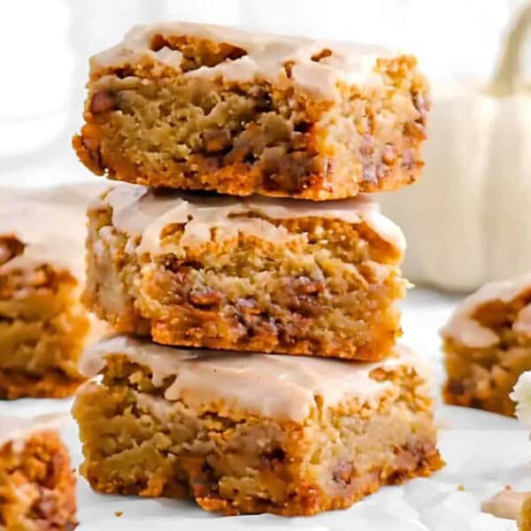 A stack of three glazed pumpkin bars is surrounded by more pumpkin bars with a white pumpkin in the background.