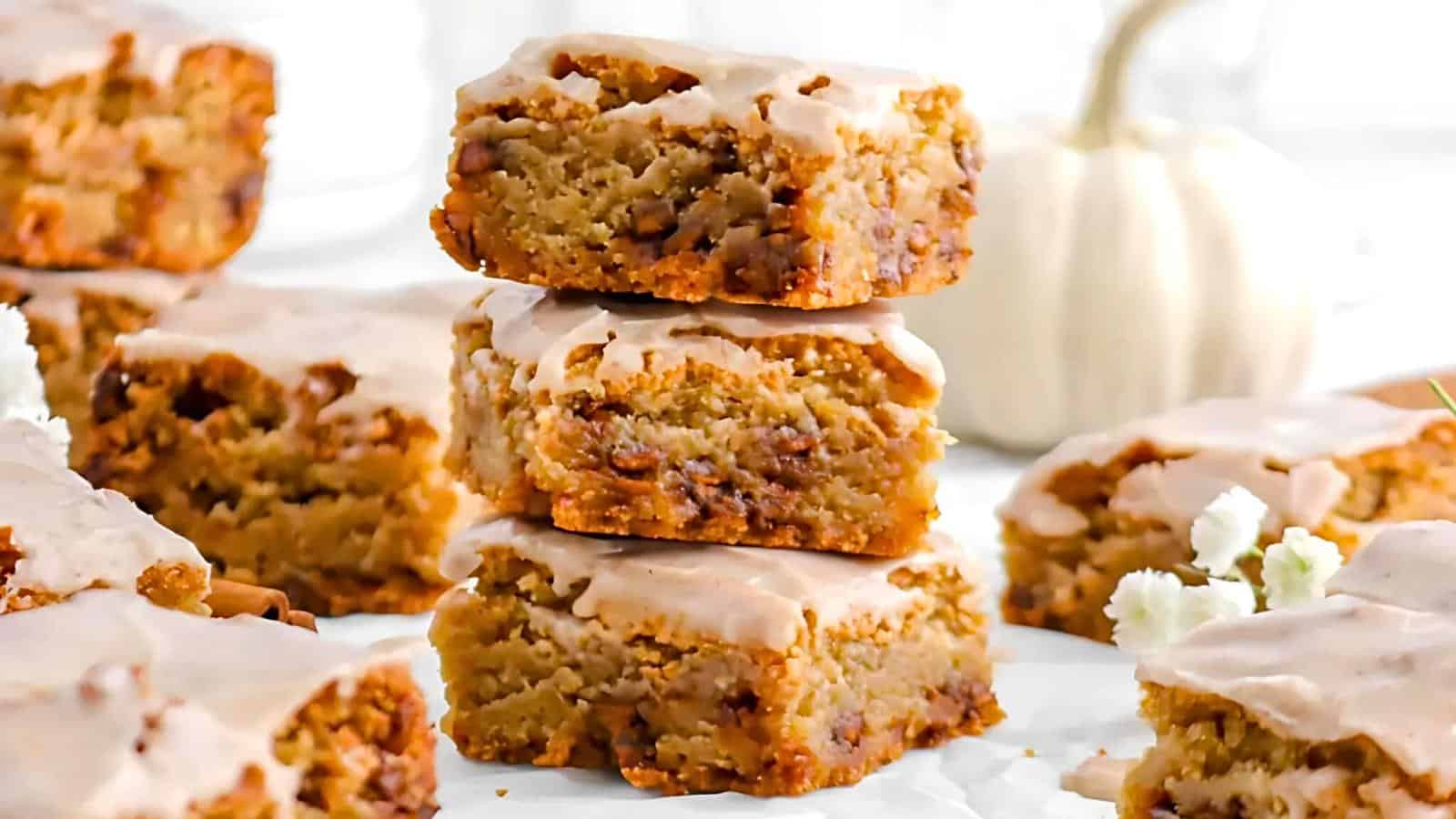 A stack of three frosted pumpkin blondies sits on parchment paper surrounded by more blondies. The blondies are golden brown with a light glaze on top. A small white pumpkin and some flowers are blurred in the background.
