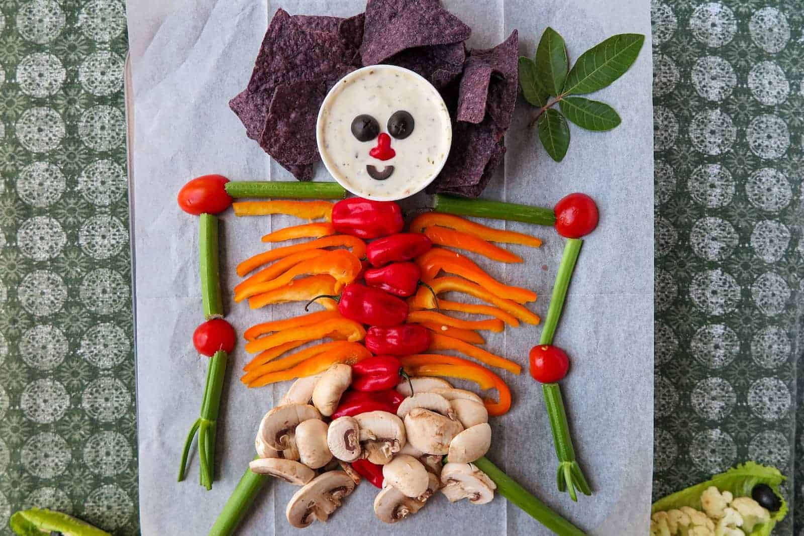 A creative food art piece where vegetables and dip are arranged to look like a human skeleton. The head is made from a dip cup with olive eyes and a tomato nose, shoulders are made from purple chips, and the body consists of various sliced vegetables.