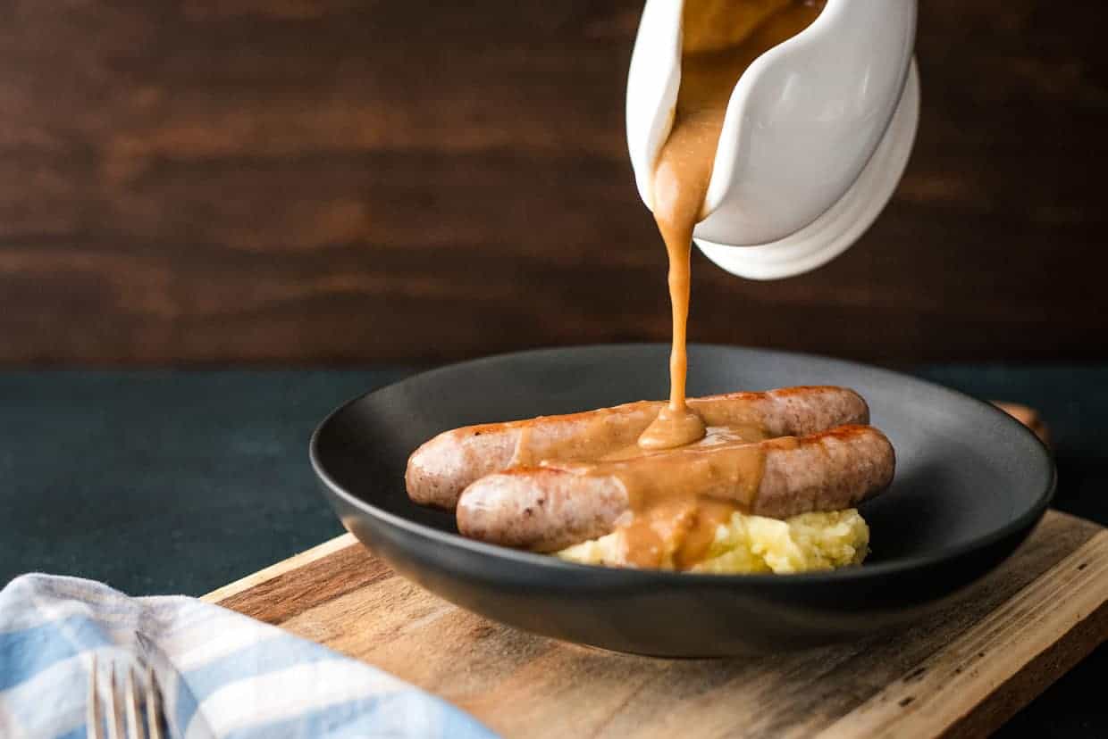 A dish of sausages served over mashed potatoes is being topped with gravy poured from a white sauce boat. The meal is presented on a black plate placed on a wooden board, with a cloth napkin nearby. The background is dark and minimalist.