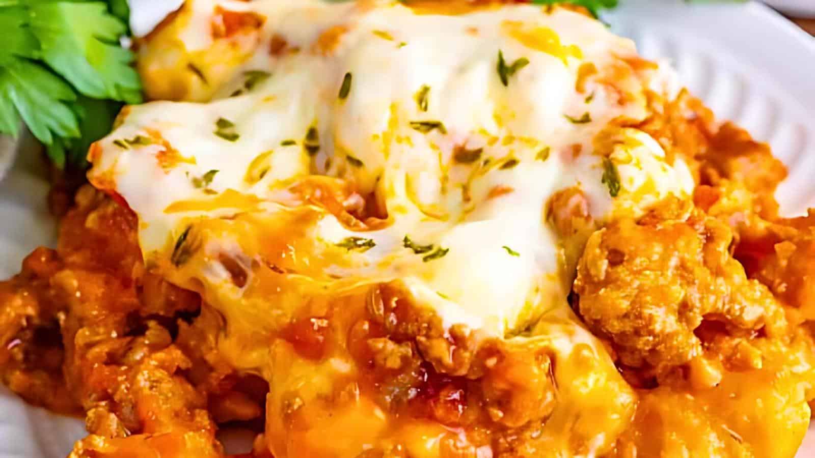 A close-up of a plate featuring cheesy baked pasta with a golden, bubbly cheese topping and a sprinkling of herbs. The dish appears creamy and savory, with chunks of meat and sauce visible beneath the melted cheese. Fresh parsley garnishes the side.