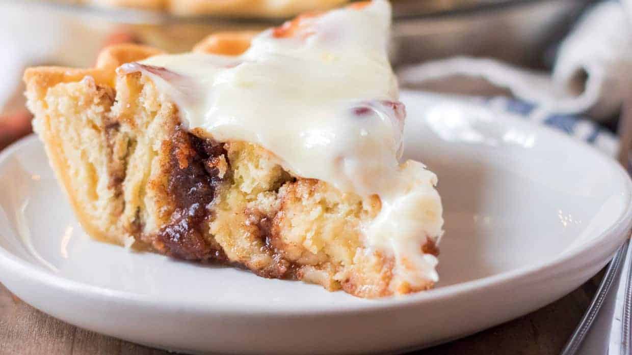 A close-up of a slice of cinnamon roll cake on a white plate. The cake is topped with a generous layer of creamy white frosting, revealing a swirl of cinnamon and sugar within the layers of the moist cake.