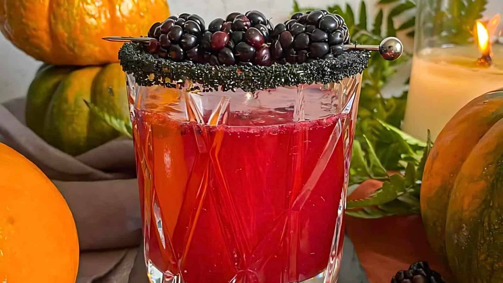 A vibrant red cocktail garnished with a skewer of blackberries and a black sugar rim sits in a faceted glass. The background includes pumpkins, green leaves, and a lit candle, adding a cozy, autumnal atmosphere.