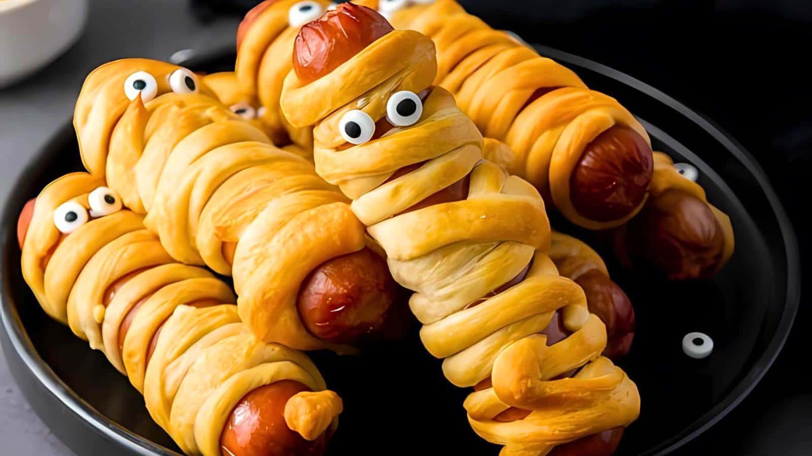 A plate of hot dogs wrapped in dough, resembling mummies, with small googly eyes placed on the dough. The hot dogs are arranged to appear playful and whimsical, perfect for a Halloween-themed treat.