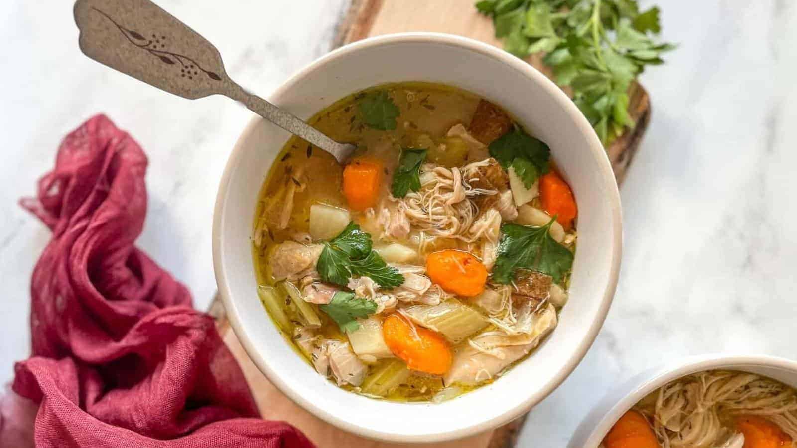 A bowl of homemade chicken soup is garnished with fresh herbs and filled with chunks of chicken, potatoes, carrots, and celery. A spoon rests in the bowl. Nearby, there is a red cloth napkin and a bunch of fresh cilantro on a cutting board.