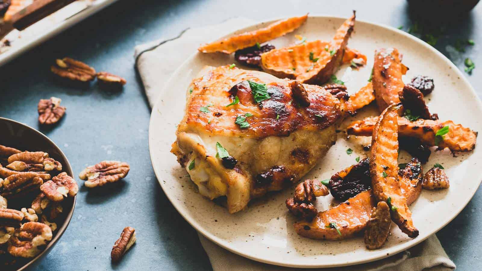 A plate with roasted chicken thigh garnished with herbs, served with sweet potato fries, pecans, and dried cranberries. Scattered pecans and a bowl of pecans are beside the plate on a gray surface.