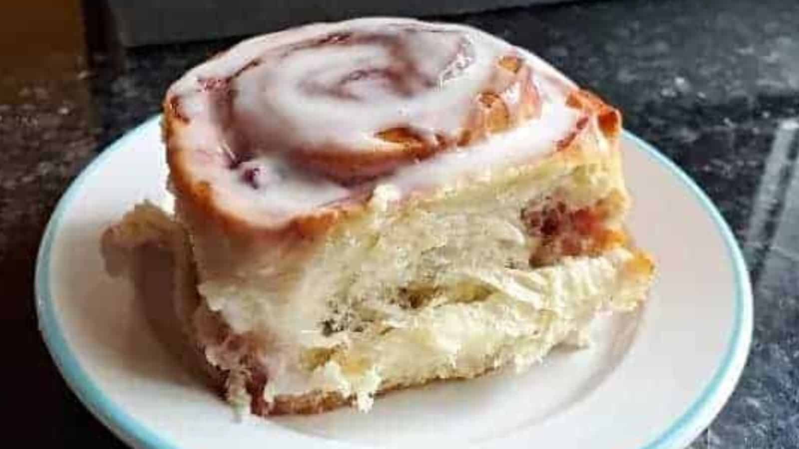 A close-up of a cinnamon roll on a white plate with a thin blue rim. The roll is topped with a generous layer of white icing and has a flaky, soft texture with visible cinnamon swirls inside. The background is a dark, speckled countertop.