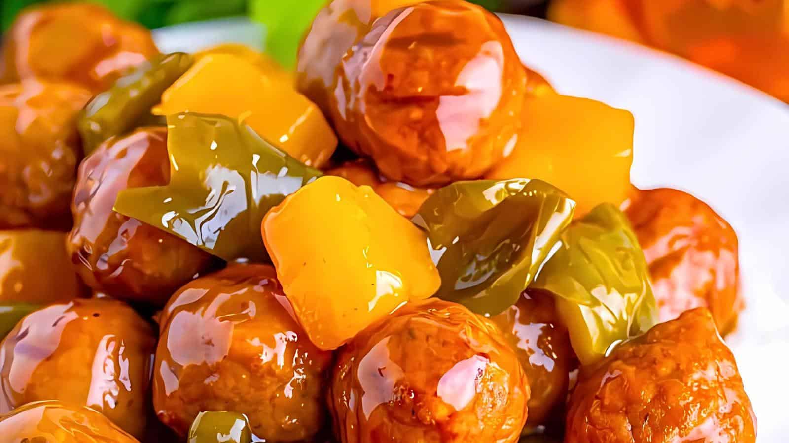 Close-up image of sweet and sour meatballs, drenched in a glossy sauce. The dish features chunks of yellow pineapple and green bell peppers, creating a vibrant, colorful presentation. The background is slightly blurred to emphasize the dish’s appetizing texture.