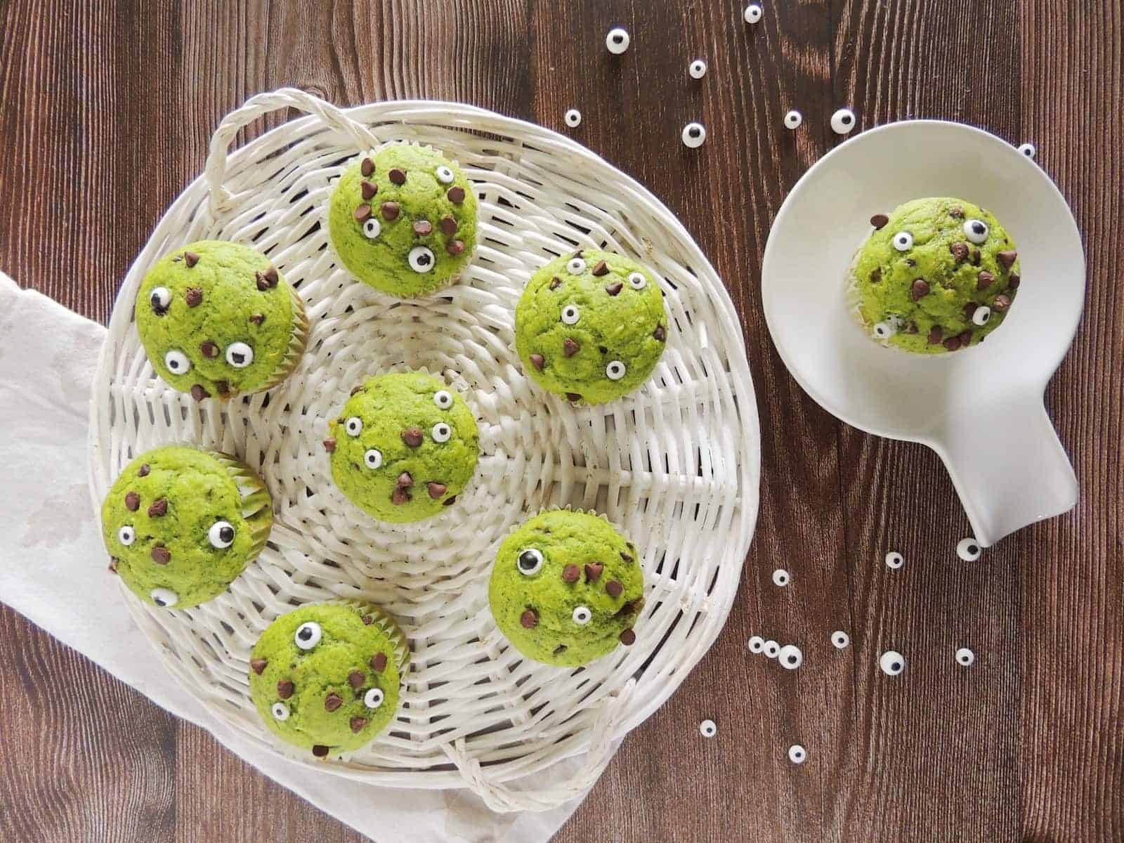 A wicker basket holds six green cupcakes decorated with candy eyes and chocolate chips, arranged on a wooden table. Left front has a white cloth. A white dish holds one additional cupcake and scattered candy eyes are around. The overall scene is whimsical and playful.