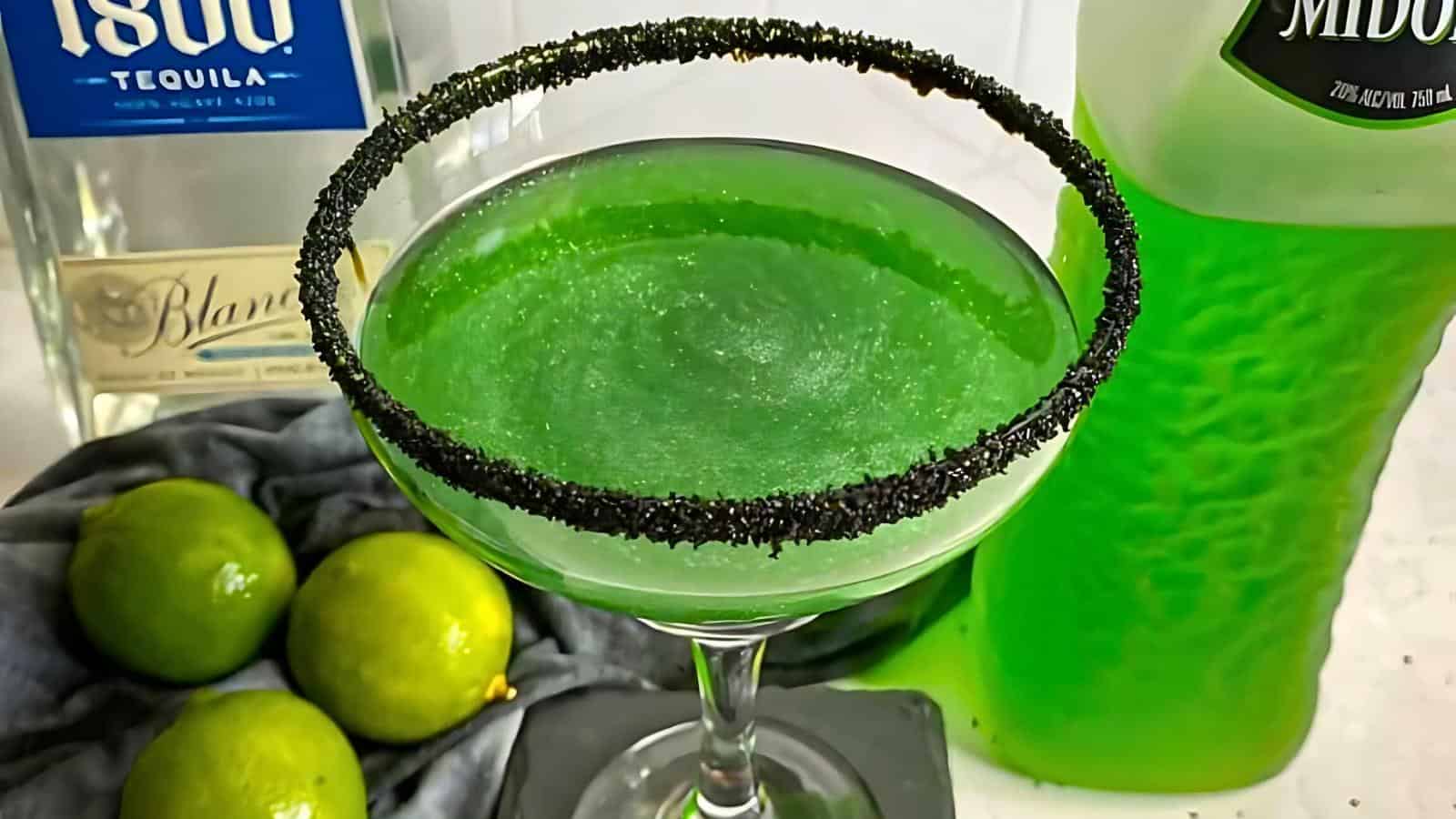 A cocktail glass with a black sugar rim sits filled with a vibrant green drink. Next to the glass is a bottle of 1800 tequila, a green bottle of Midori, and a few fresh limes, all arranged on a white surface.