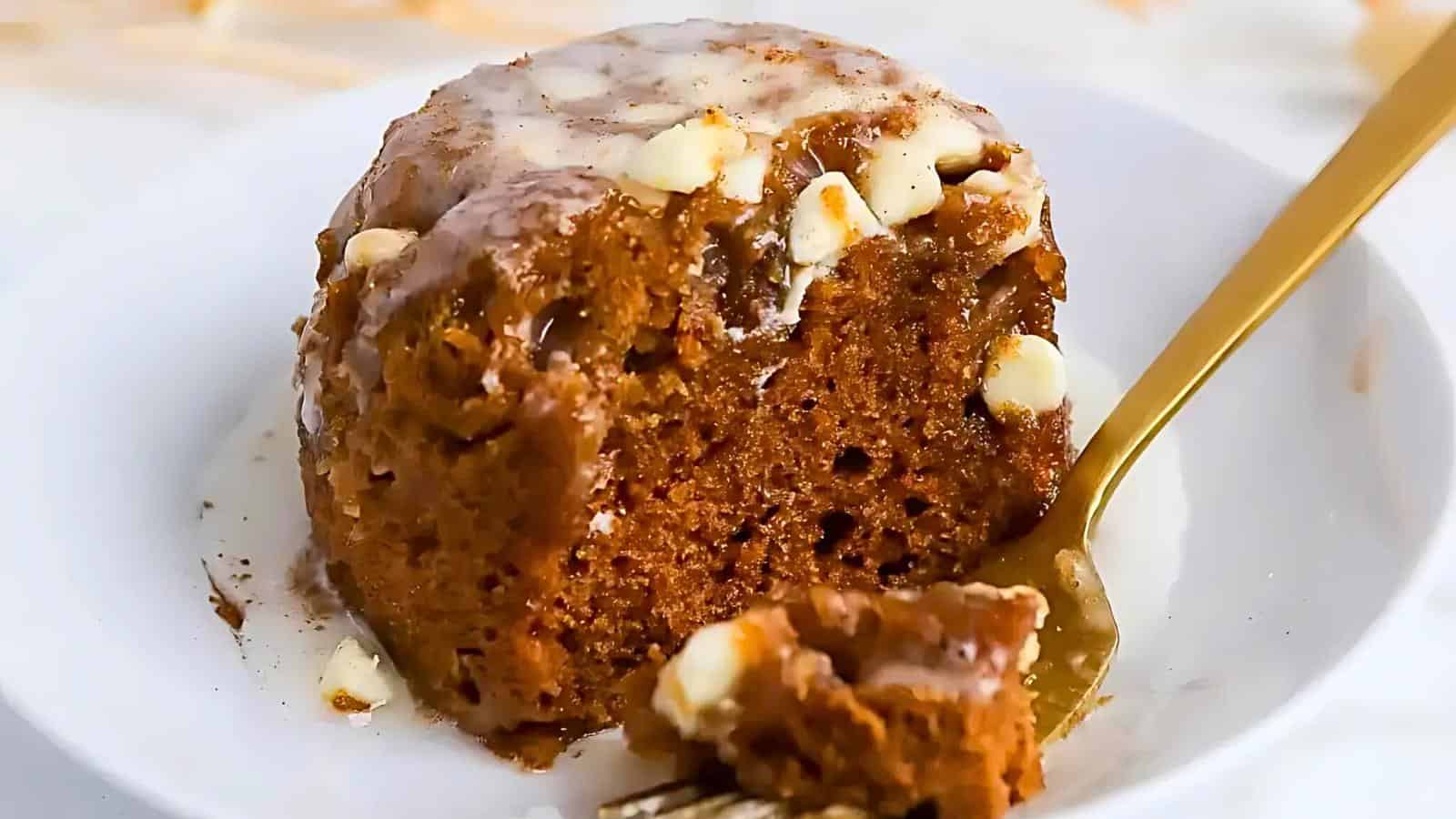 A close-up of a delicious, gooey cinnamon roll cake on a white plate. It's topped with a drizzle of white icing and sprinkled with white chocolate chips. A bite has been taken out, and a gold fork with a piece of cake on it is resting on the plate.