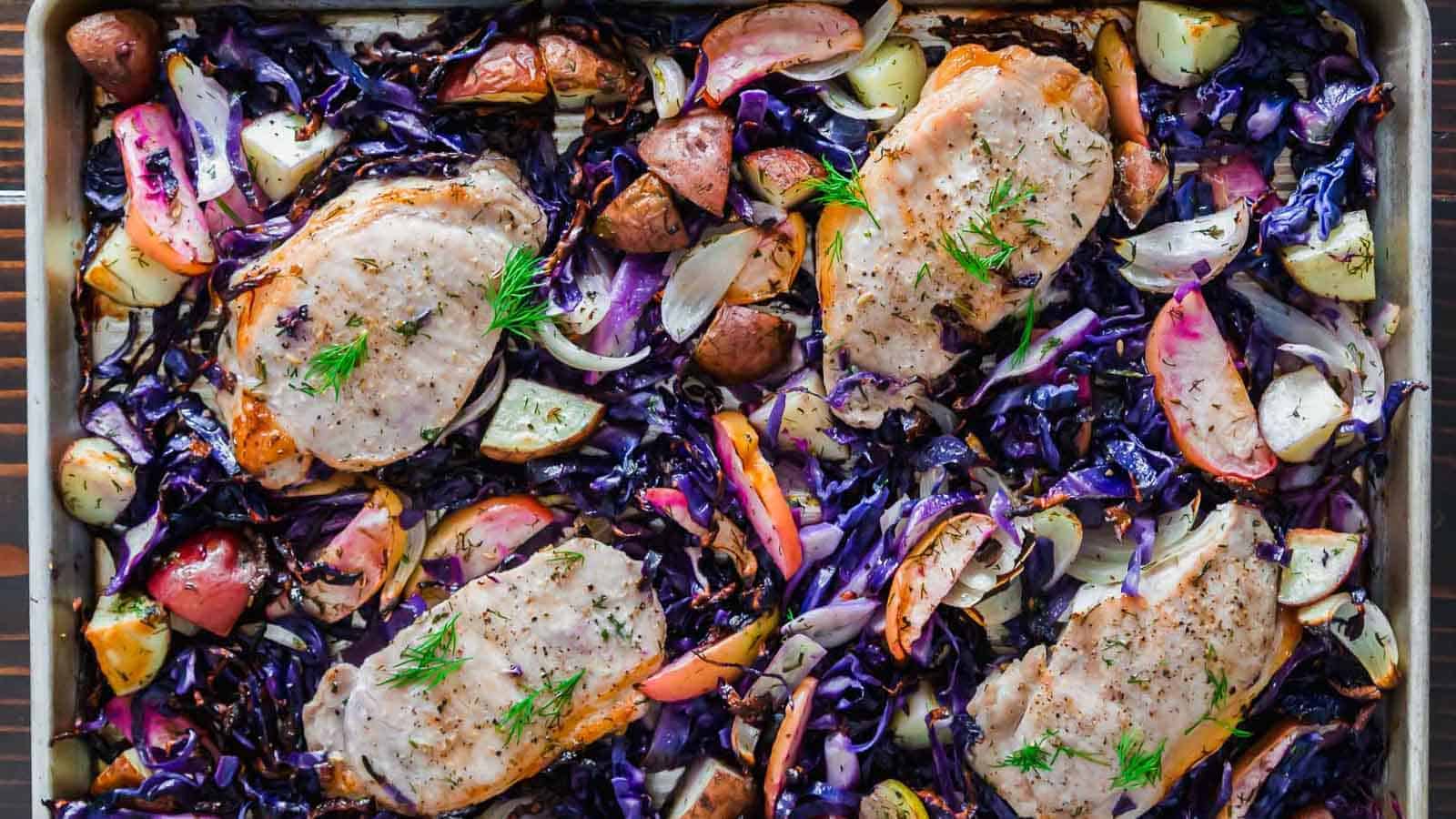 A baking sheet filled with roasted pork chops, sliced red cabbage, apples, red potatoes, and onion slices, all seasoned and garnished with sprigs of fresh herbs. The colorful assortment creates a vibrant and appetizing display.