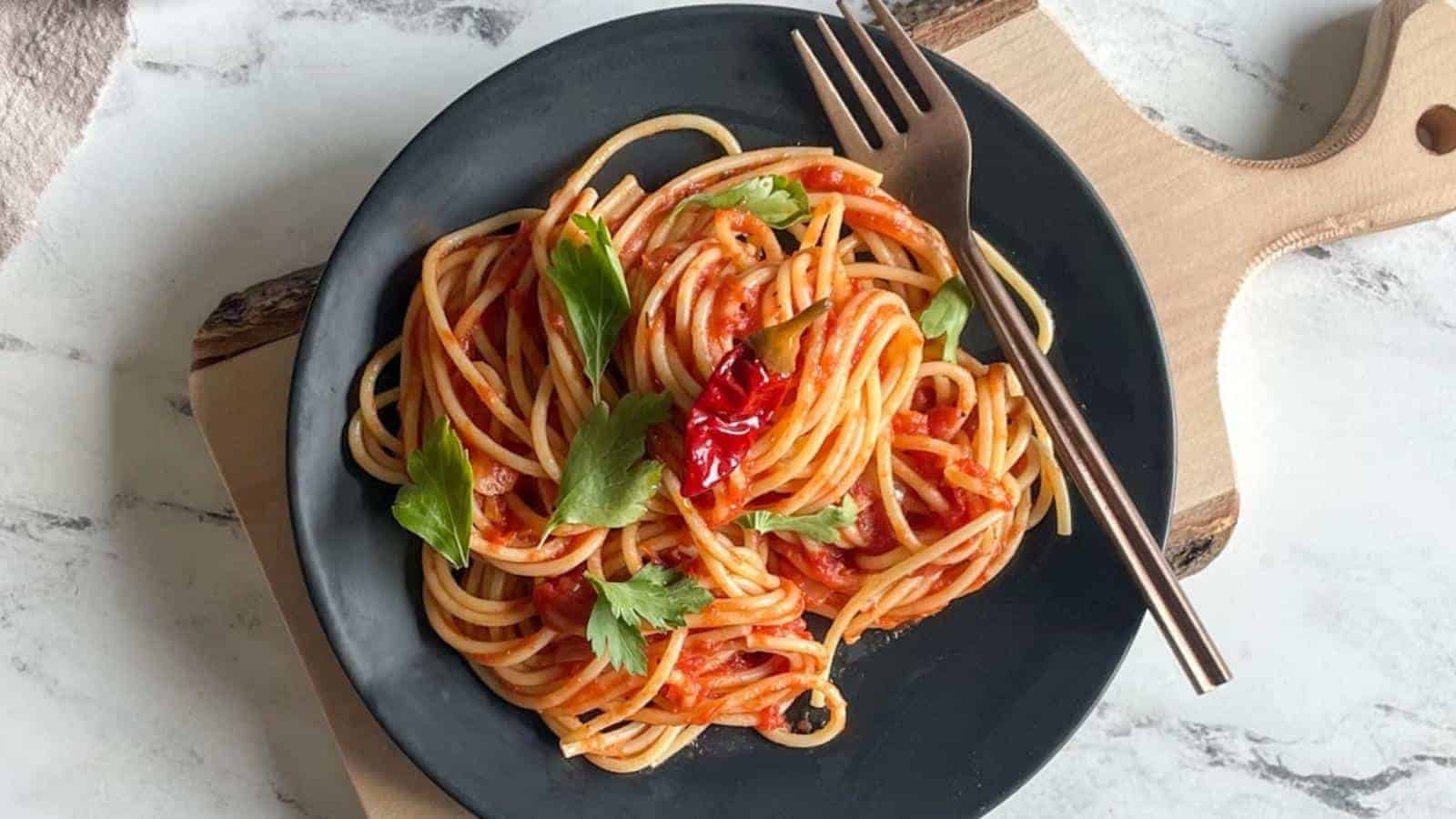 A black plate filled with spaghetti topped with tomato sauce, garnished with fresh parsley and red chili peppers. A fork rests on the side of the plate, which is placed on a wooden cutting board on a marble surface.