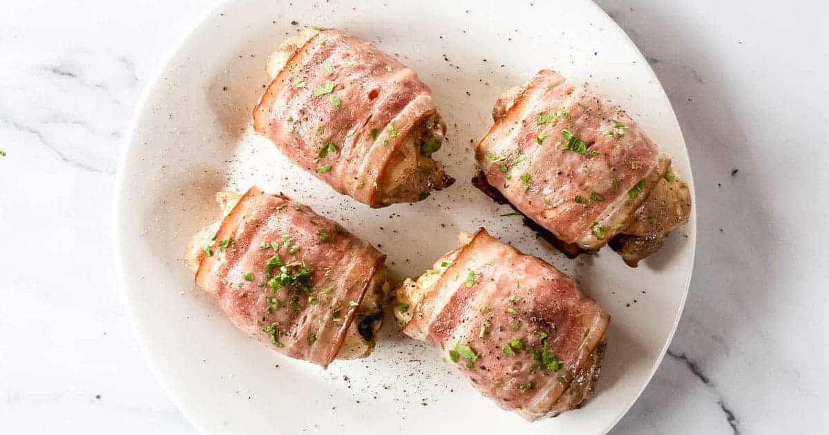 A white plate with four bacon-wrapped chicken breasts garnished with finely chopped herbs. The chicken rolls are crispy and golden, and the plate sits on a light-colored marble surface.