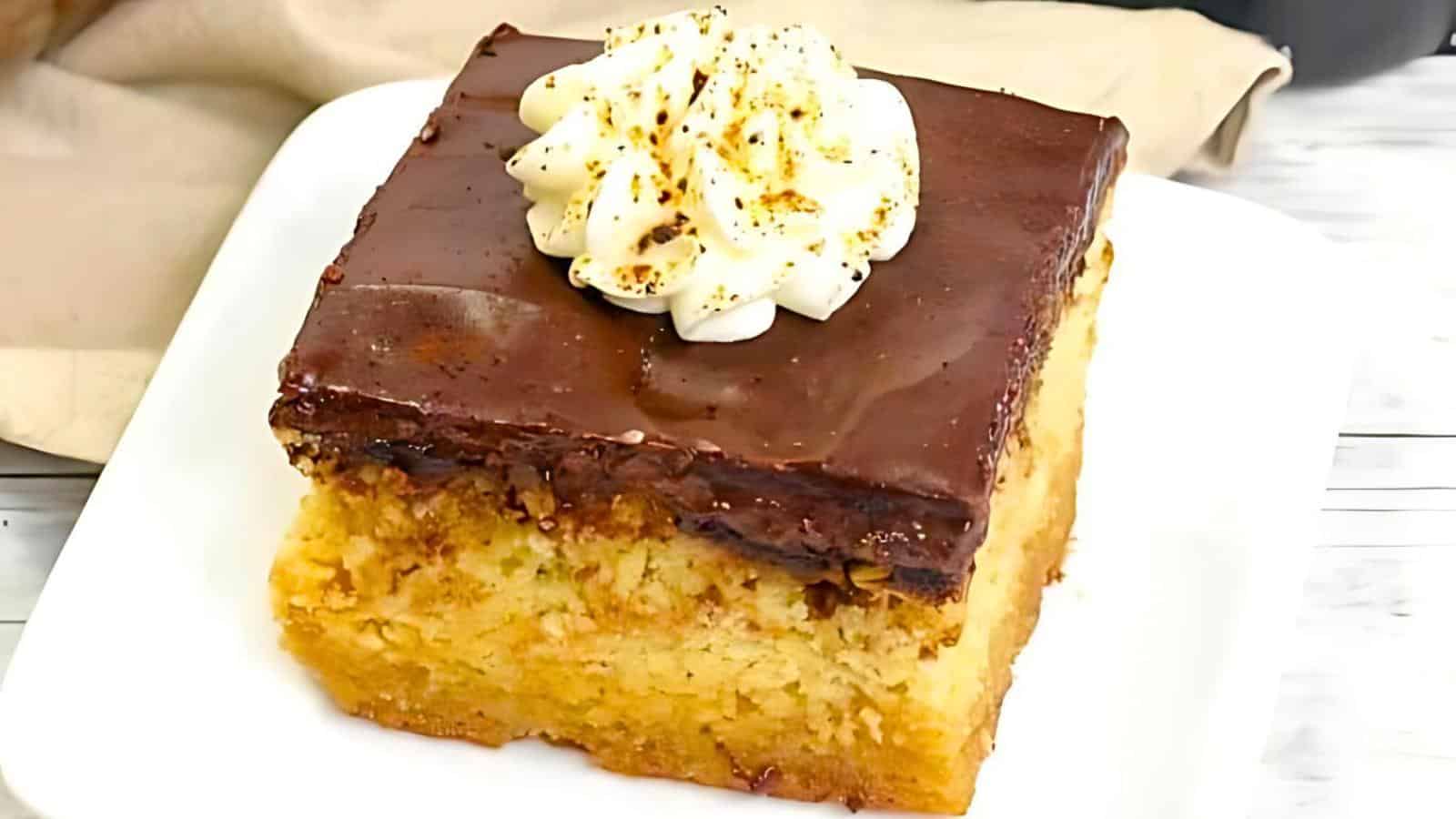 A slice of cake on a white plate. The cake has a yellow base with a crumbly texture, topped with a layer of rich chocolate glaze. A dollop of whipped cream with a sprinkle of cocoa powder sits on top. The background is a neutral-toned fabric.