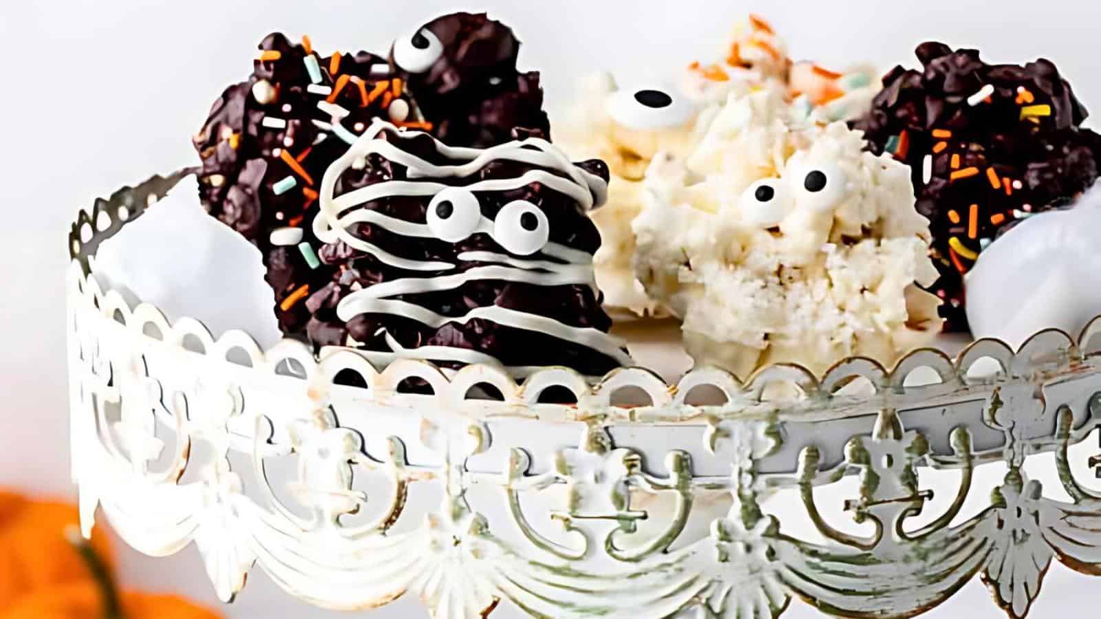 A decorative metal stand holds assorted Halloween-themed treats. The treats are chocolate-covered clusters adorned with candy eyes and festive sprinkles, resembling monsters or creepy faces. They come in both dark and white chocolate varieties.