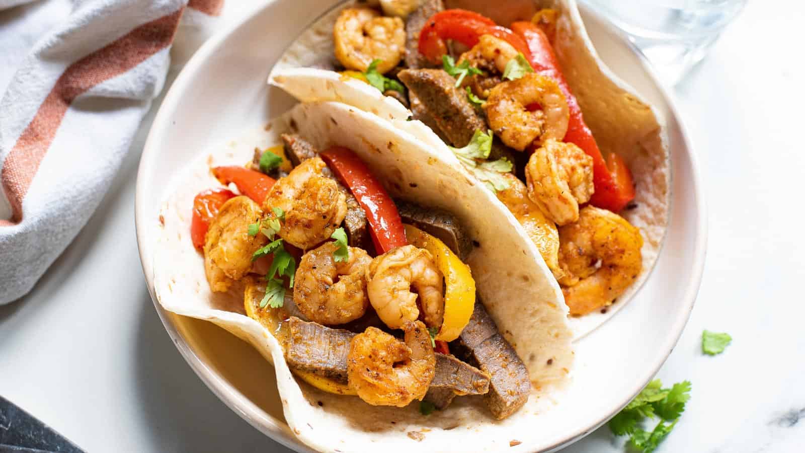 A plate of shrimp fajitas featuring two tortillas filled with seasoned shrimp, red and yellow bell peppers, and strips of steak, garnished with cilantro. A glass of water and a napkin are visible nearby.