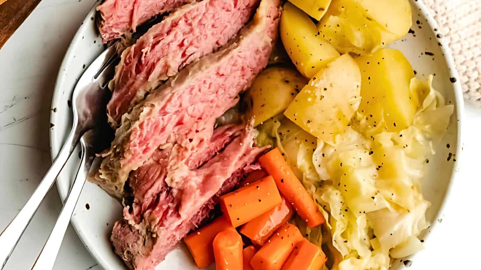 A plate filled with sliced corned beef, boiled potatoes, carrots, and cabbage. The corned beef is pink and tender, the potatoes are yellow, and the carrots are orange. Ground pepper is sprinkled on top of the vegetables, and there is a fork and spoon on the side.