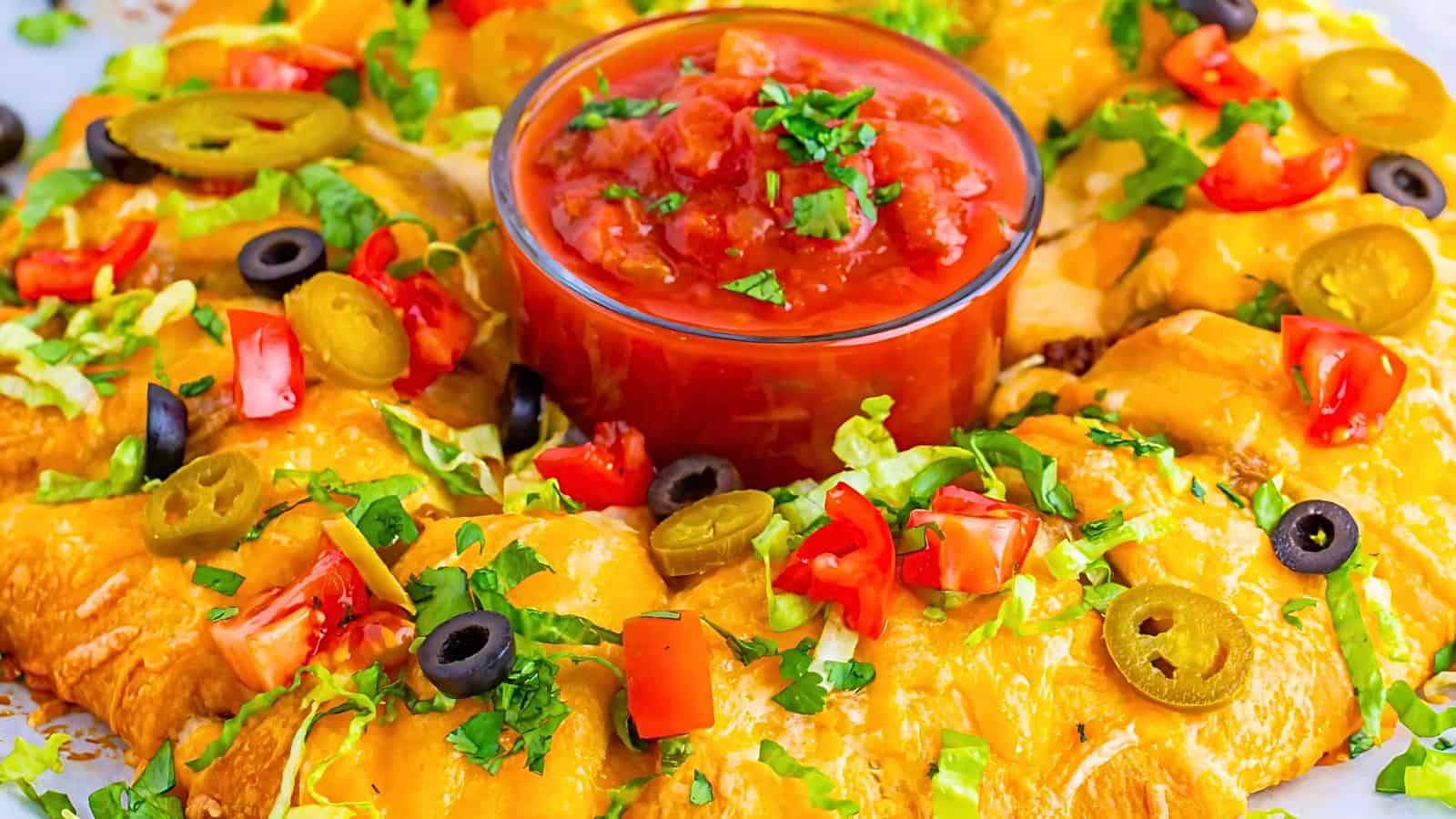 A colorful plate of cheesy baked crescent rolls arranged in a circular pattern, topped with chopped tomatoes, black olives, jalapeños, shredded lettuce, and fresh cilantro. In the center, a bowl filled with red salsa garnished with green herbs completes the vibrant dish.