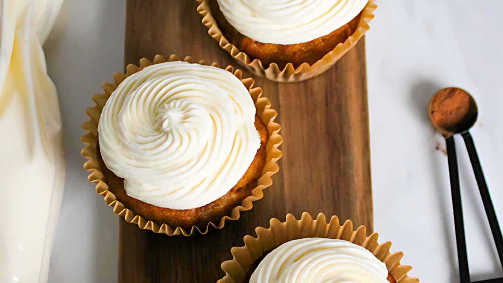 Three cupcakes with swirled white frosting sit on a wooden board. A small metal measuring spoon with a pinch of cinnamon is nearby. The cupcakes are in golden brown paper liners and arranged neatly, creating a visually appealing presentation.