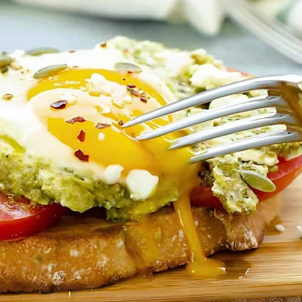A fork breaking the yolk of a poached egg on avocado toast with tomato slices and sprinkled feta cheese on a wooden serving board.