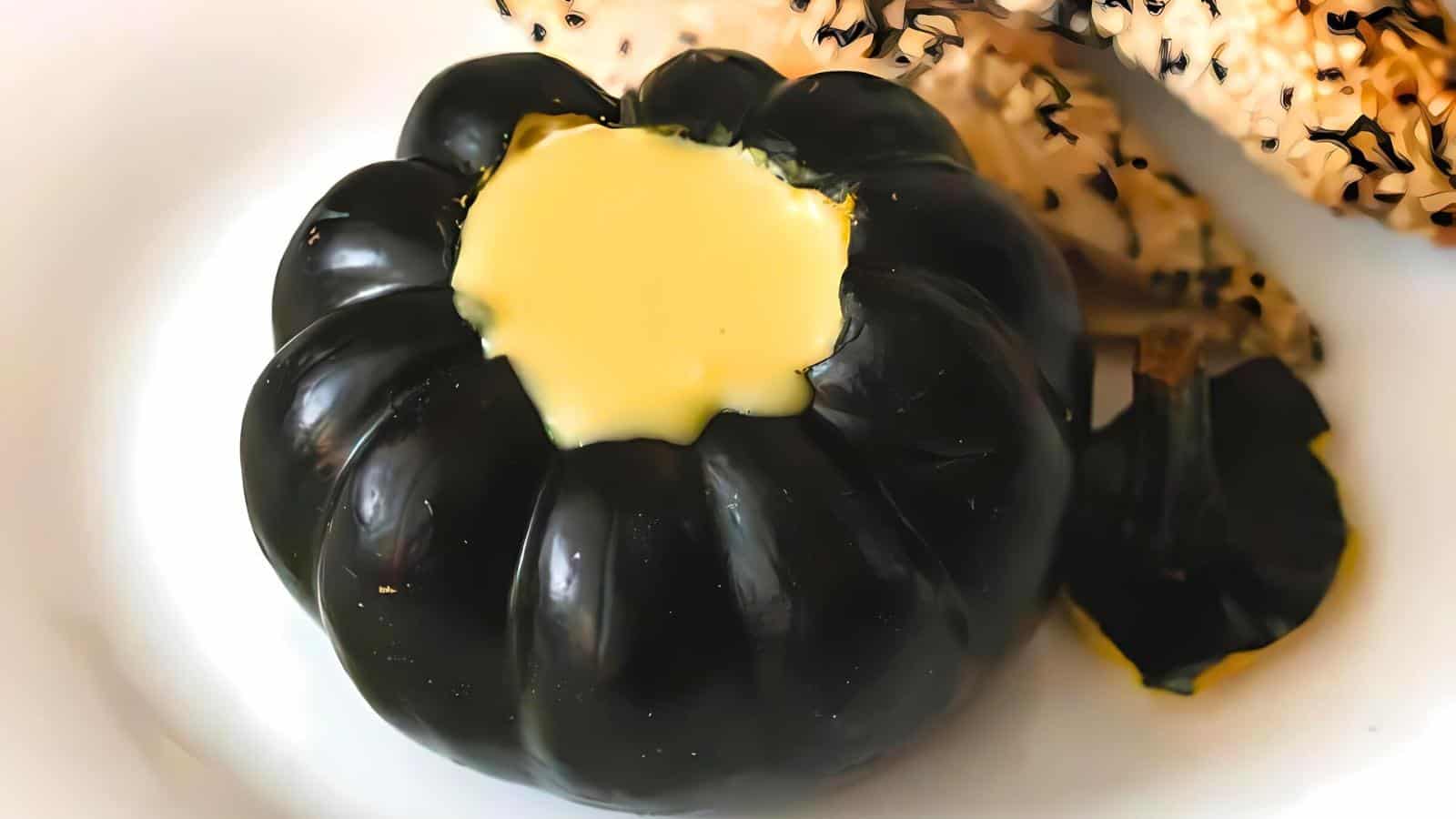 A small black pumpkin filled with creamy yellow custard sits on a white plate. The pumpkin has smooth, shiny skin and ridges. In the background, there are some brown elements possibly resembling toasted bread or crackers.