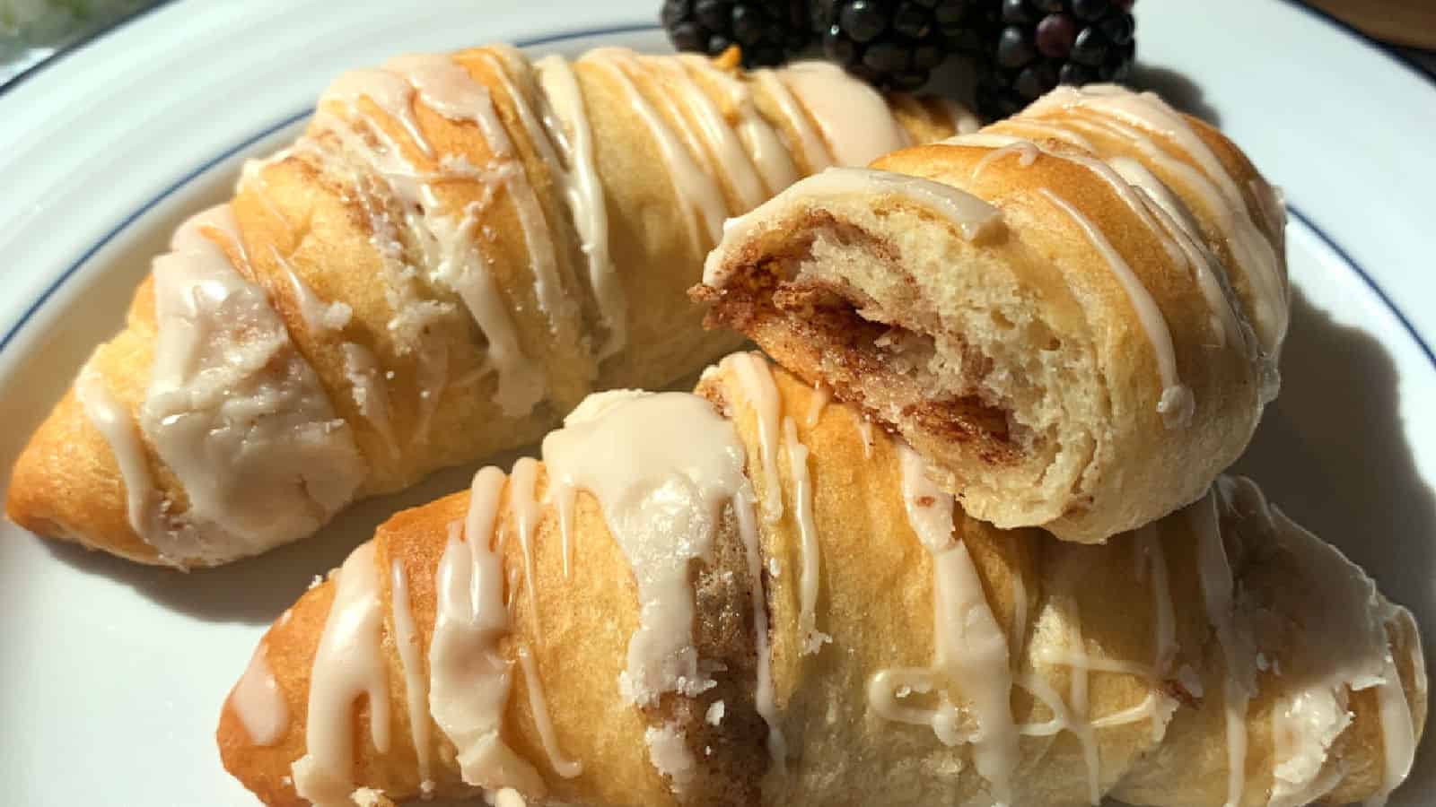 Two golden-brown crescent rolls drizzled with white icing are placed on a white plate with a black and blue trim. One roll is partially eaten, revealing a cinnamon filling. A few blackberries are positioned in the background.