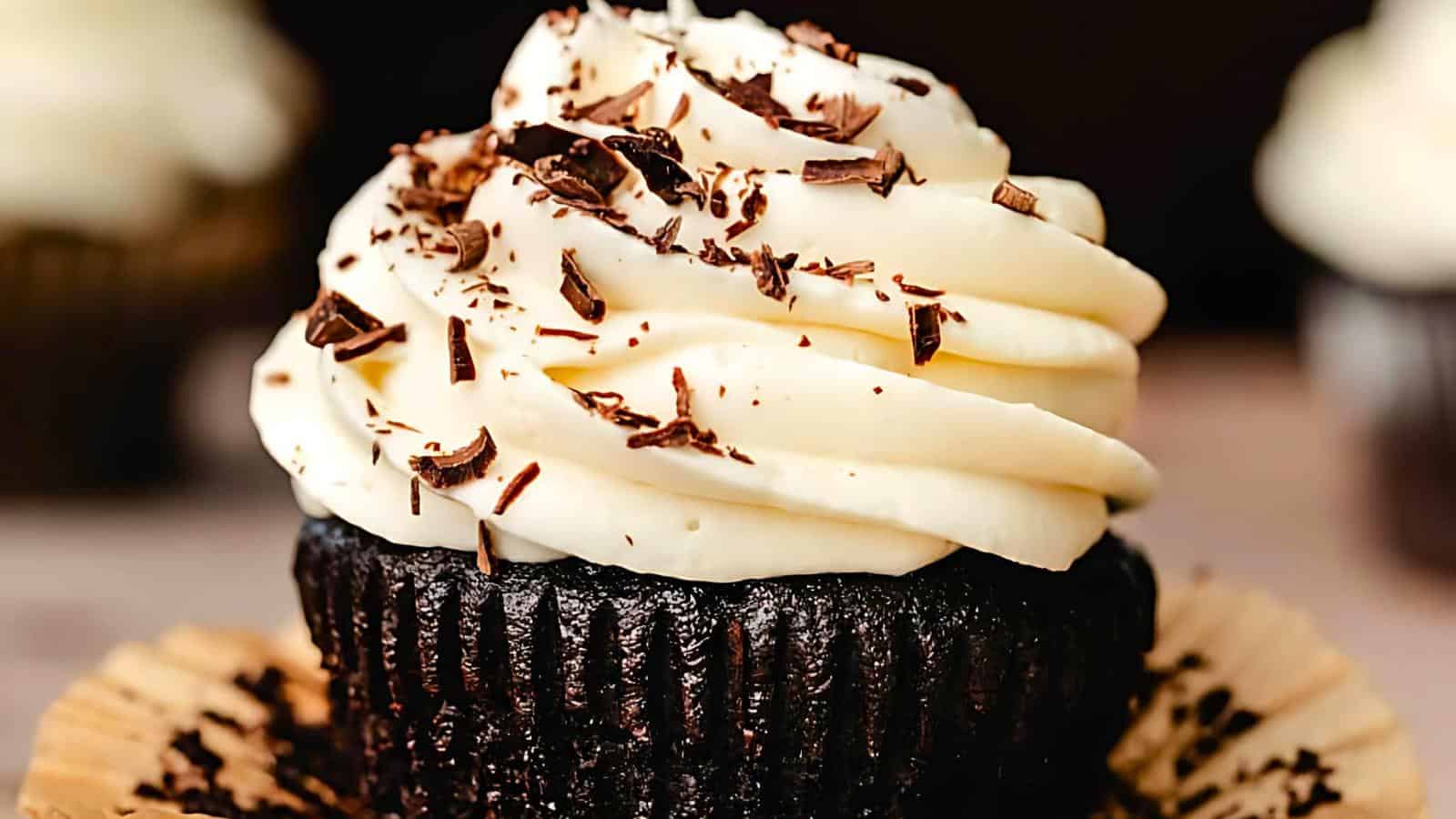 A close-up of a chocolate cupcake with a swirl of creamy white frosting, topped with chocolate shavings. The cupcake is partially unwrapped from its brown paper liner, showcasing its rich texture and enticing appearance.
