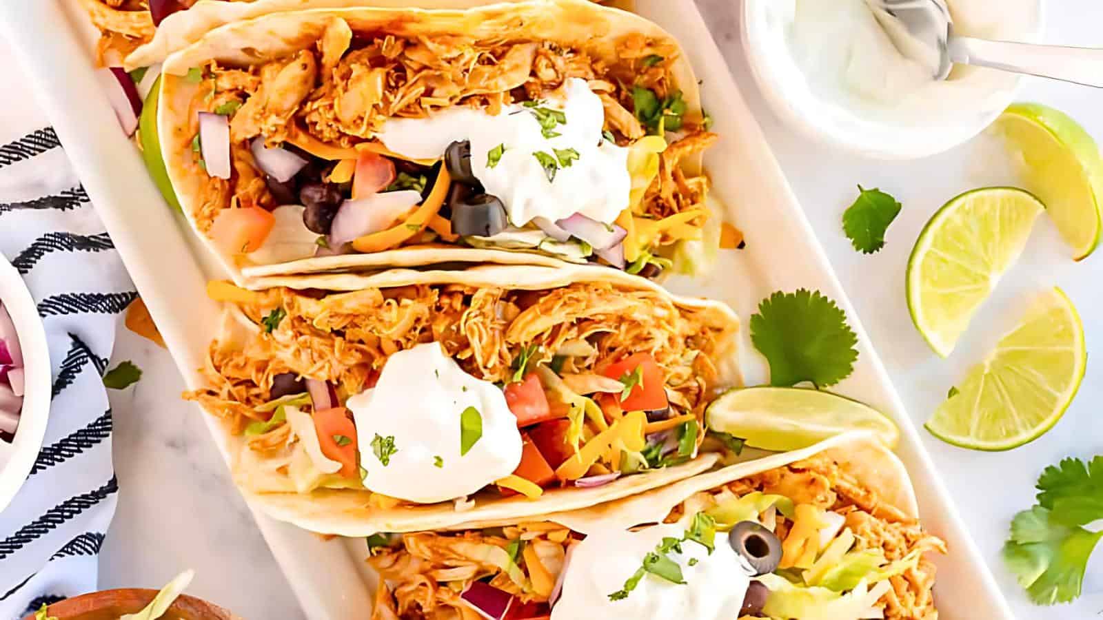 A close-up of three tacos filled with shredded chicken, diced tomatoes, onions, olives, lettuce, and topped with dollops of sour cream. The tacos are garnished with fresh cilantro and served with lime wedges on the side.
