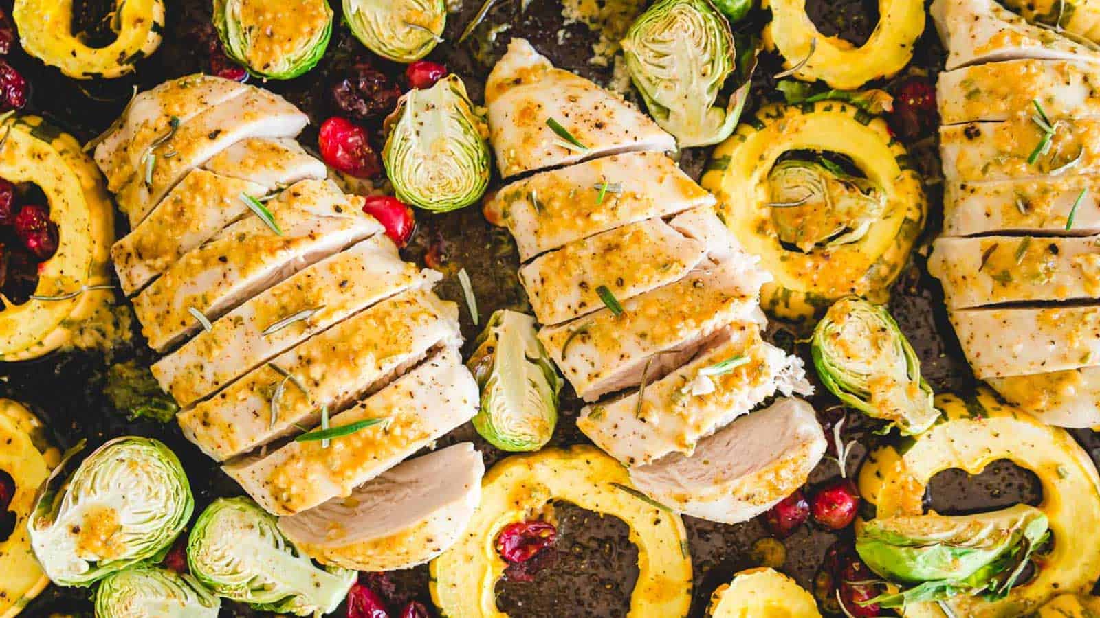A close-up of a baked chicken dish featuring sliced chicken breasts garnished with herbs. Surrounding the chicken are roasted Brussels sprouts, cranberries, and sliced winter squash, all placed on a baking sheet. The dish is seasoned and perfectly browned.