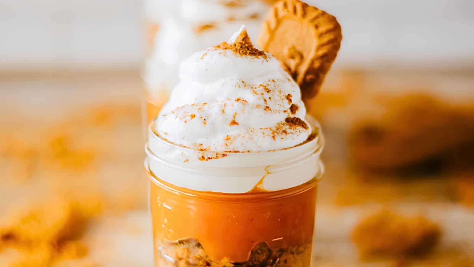 A dessert jar filled with pumpkin pie filling, topped with whipped cream, a sprinkle of spices, and a cookie garnish. The jar has layered crumbs at the bottom, and the background features a blurred view with scattered crumbs and additional desserts.