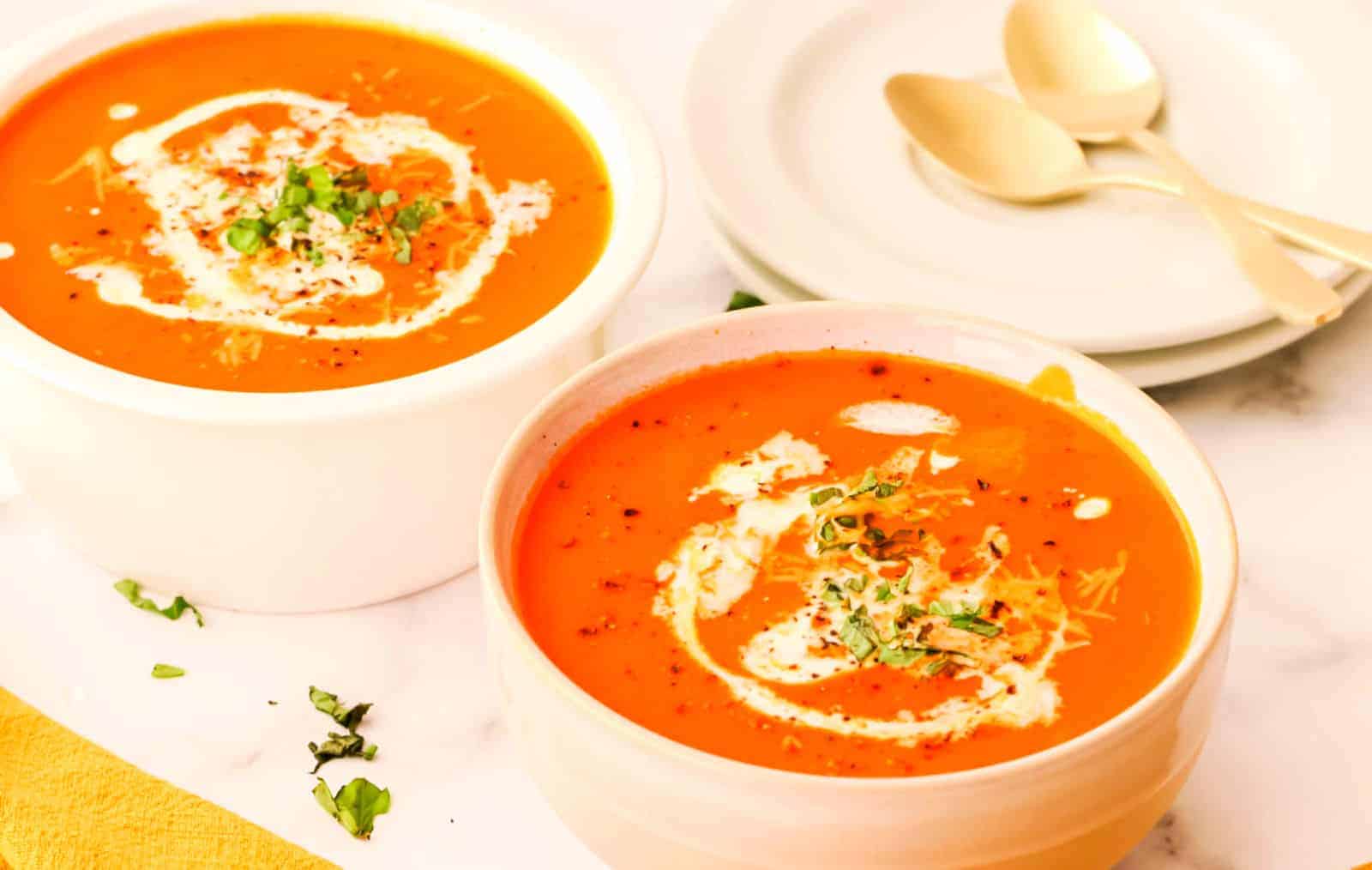 Two bowls of creamy tomato soup are garnished with a swirl of cream and fresh herbs. The bowls are placed on a light-colored surface next to an empty plate with two gold-colored spoons on it.