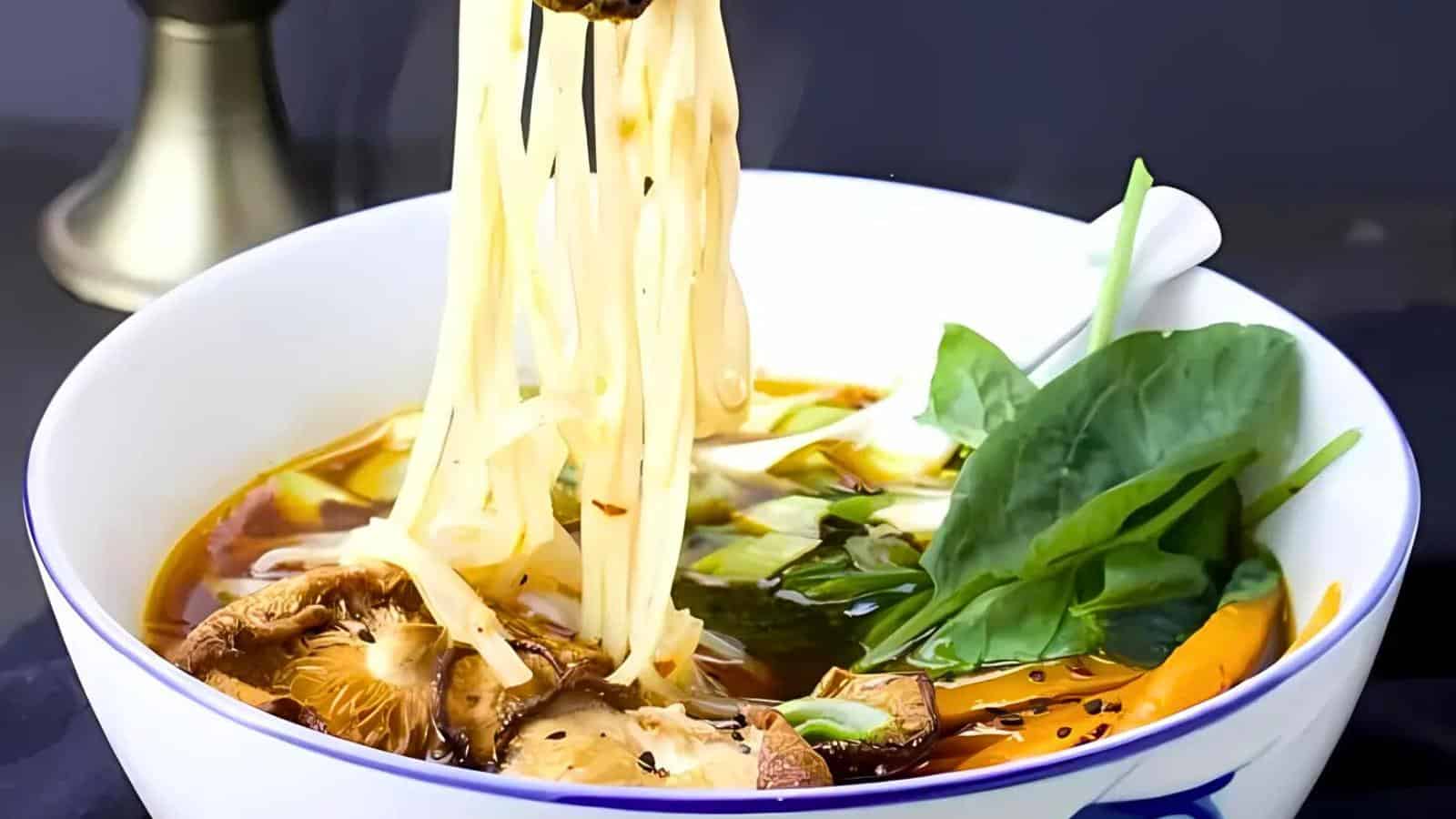 A bowl of ramen noodle soup with shiitake mushrooms, leafy greens, and other vegetables. Chopsticks are lifting noodles from the broth, which appears rich and flavorful. A white spoon rests in the bowl, ready for eating.