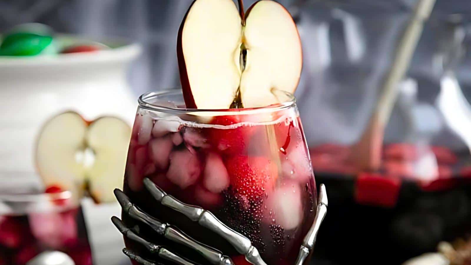 A glass filled with a red beverage and ice, garnished with two apple slices forming the shape of a heart at the rim. The glass is held by a decorative skeletal hand, adding a touch of spookiness. In the background, blurred out, are other drinks and a bowl.
