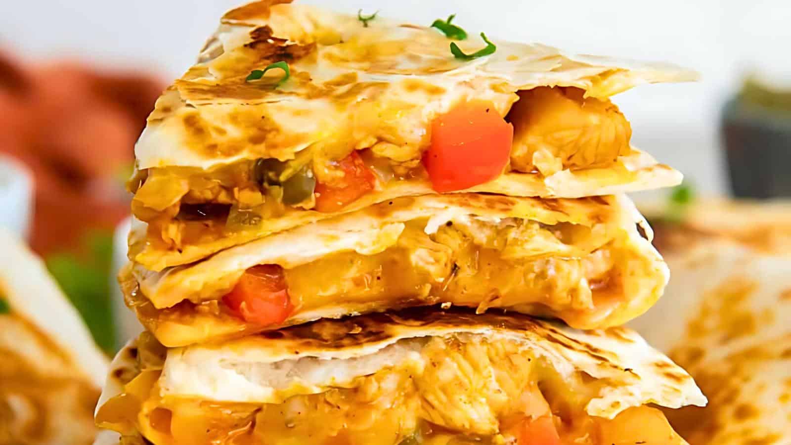 Close-up of a stack of cheesy quesadillas filled with melted cheese, diced tomatoes, green peppers, and bits of chicken. The tortillas are golden brown and slightly crispy, garnished with a few sprigs of green herbs on top.