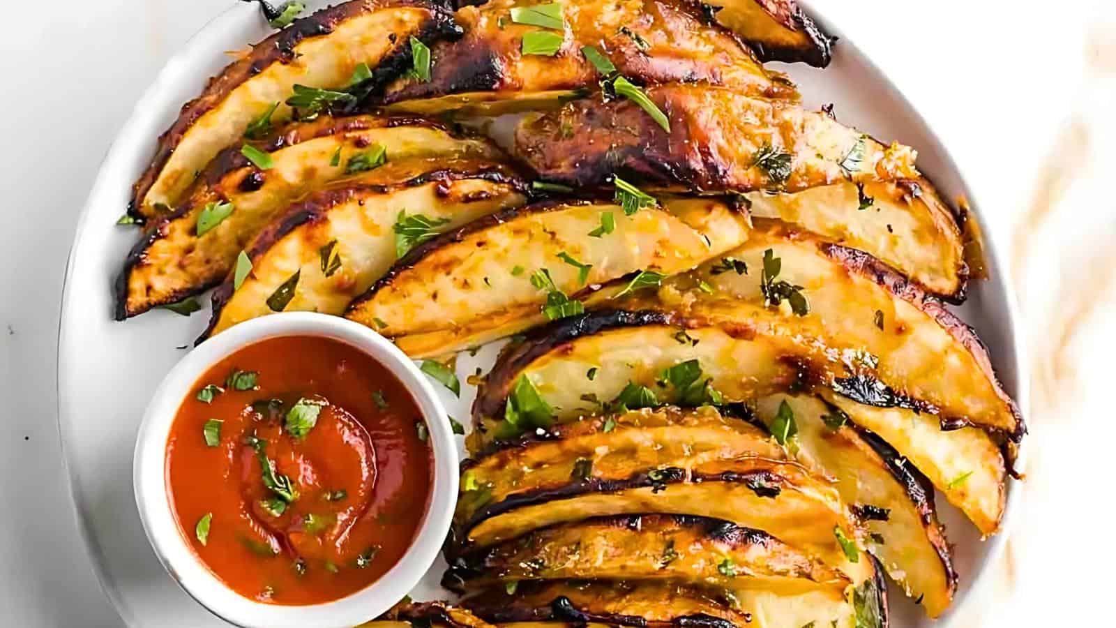 A plate of grilled potato wedges garnished with chopped parsley is arranged in a circular pattern. A small white bowl filled with red dipping sauce sits on the edge of the plate. The potatoes are lightly charred and seasoned.