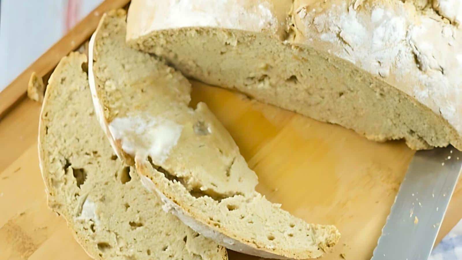 Close-up of a freshly baked loaf of bread on a wooden cutting board, with two thick slices buttered on top. A serrated knife lies nearby. The bread has a light, airy crumb and a slightly crusty exterior.