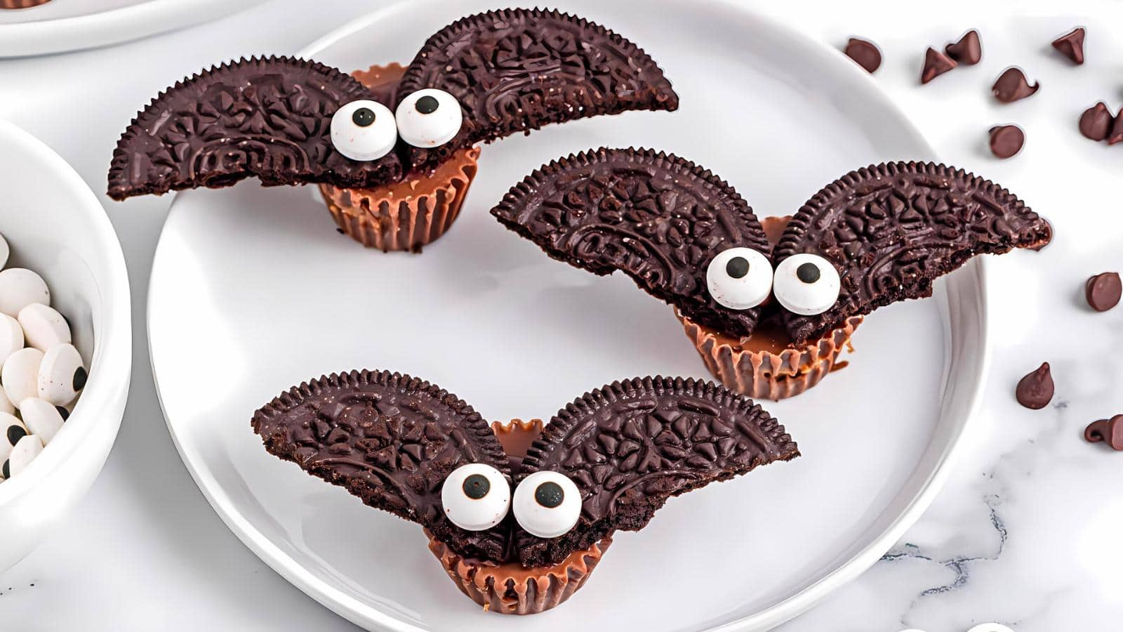 Three chocolate bat-themed desserts are displayed on a white plate. Each bat is crafted using a peanut butter cup for the body, Oreo cookie wings, and candy eyes. Chocolate chips are scattered nearby, adding to the Halloween-themed presentation.