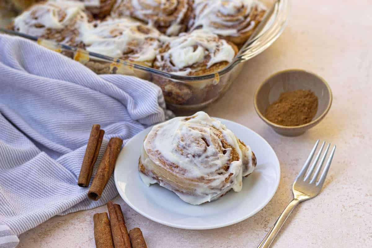 A plate with a frosted cinnamon roll sits beside a fork, several cinnamon sticks, a bowl of cinnamon, and a striped cloth. In the background, a glass baking dish holds more frosted cinnamon rolls. The scene is set on a light-colored surface.
