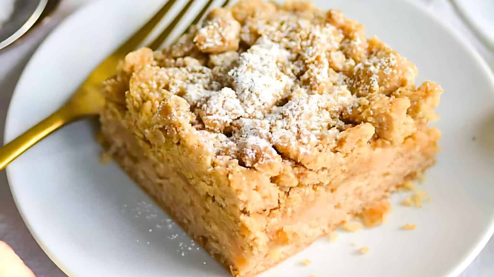 A slice of crumbly coffee cake topped with powdered sugar sits on a white plate. A gold fork accompanies the dessert, positioned to the left of the cake. The background includes partial views of additional tableware.