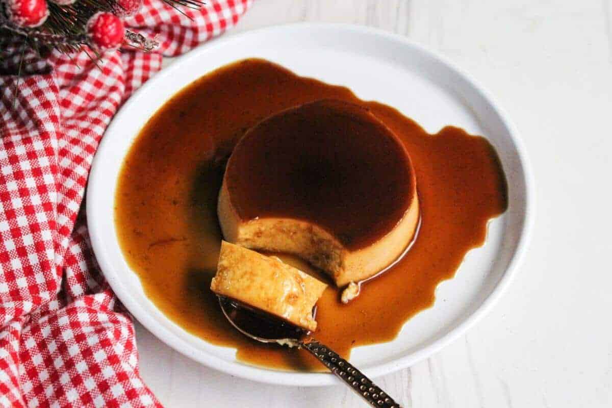 A plate of caramel flan with a piece cut out on a spoon, lying in a pool of rich caramel sauce. The plate is set on a white surface with a red and white checkered cloth nearby, adorned with small festive decorations.