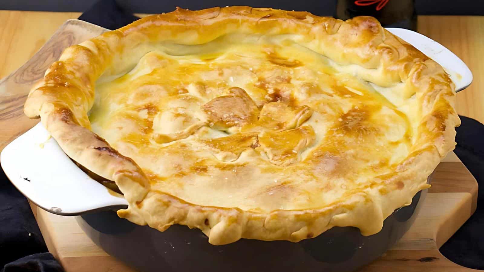 A freshly baked, golden-brown chicken pot pie in a white ceramic dish sits on a wooden surface. The pie has a flaky, slightly puffed crust with crimped edges, and the filling is not visible as it is covered completely by the crust.