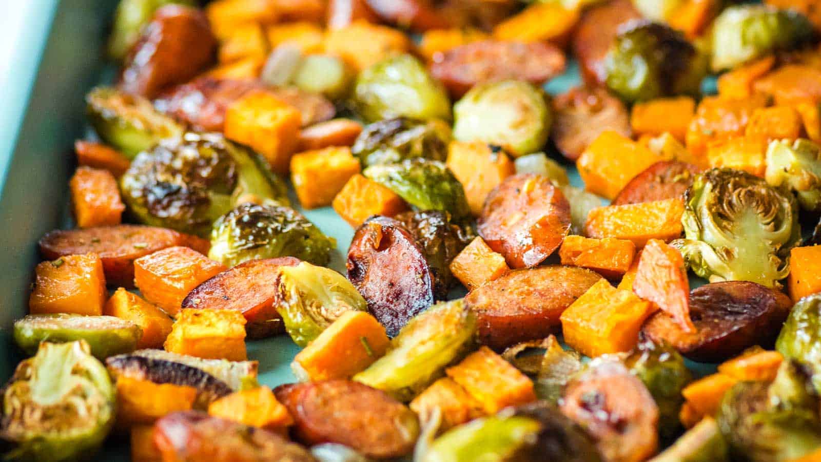 A close-up of a colorful roasted vegetable medley on a baking sheet, featuring browned brussels sprouts, vibrant orange sweet potatoes, and caramelized slices of sausage. The vegetables and sausage are evenly spread out and appear well-cooked and lightly charred.