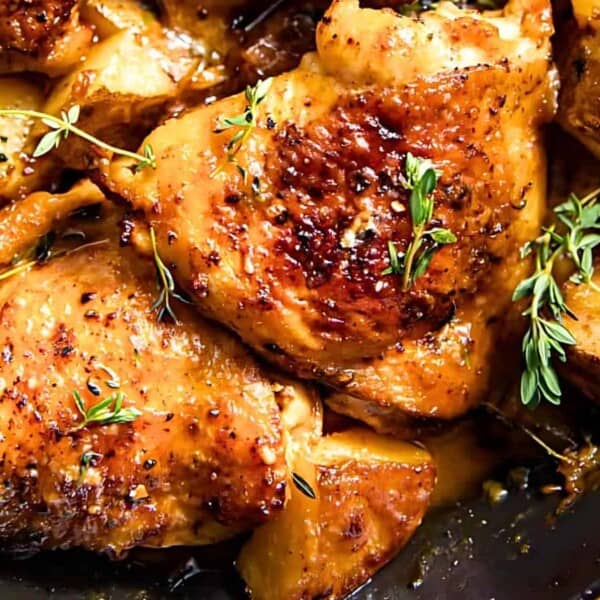 Roasted chicken thighs with crispy skin, garnished with fresh herbs, accompanied by roasted potatoes in a black skillet.
