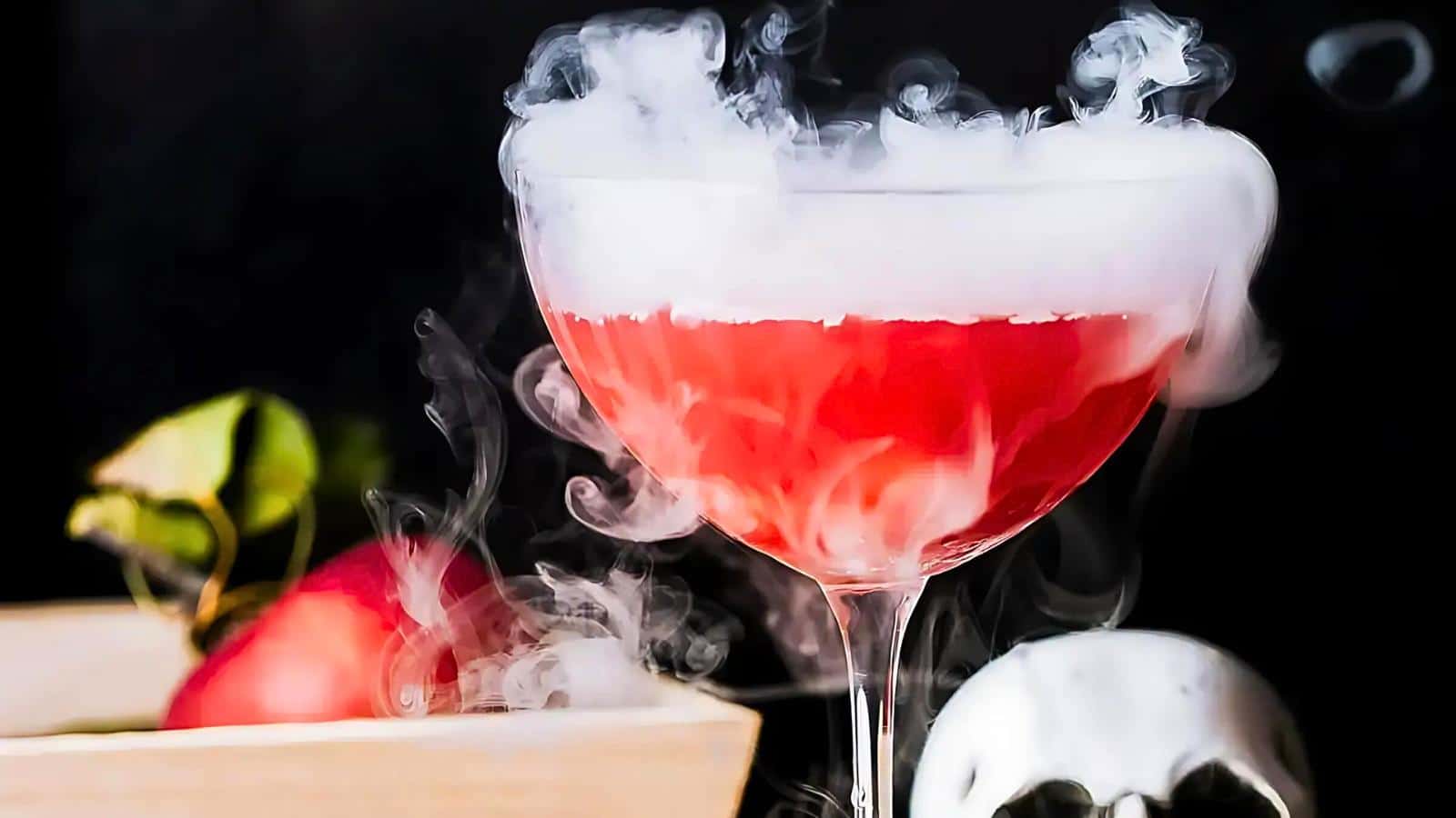 A cocktail glass filled with a red drink emits a dramatic fog, adding a mysterious and enchanting effect. In the background, there is an apple and a small white skull, all set against a dark backdrop.