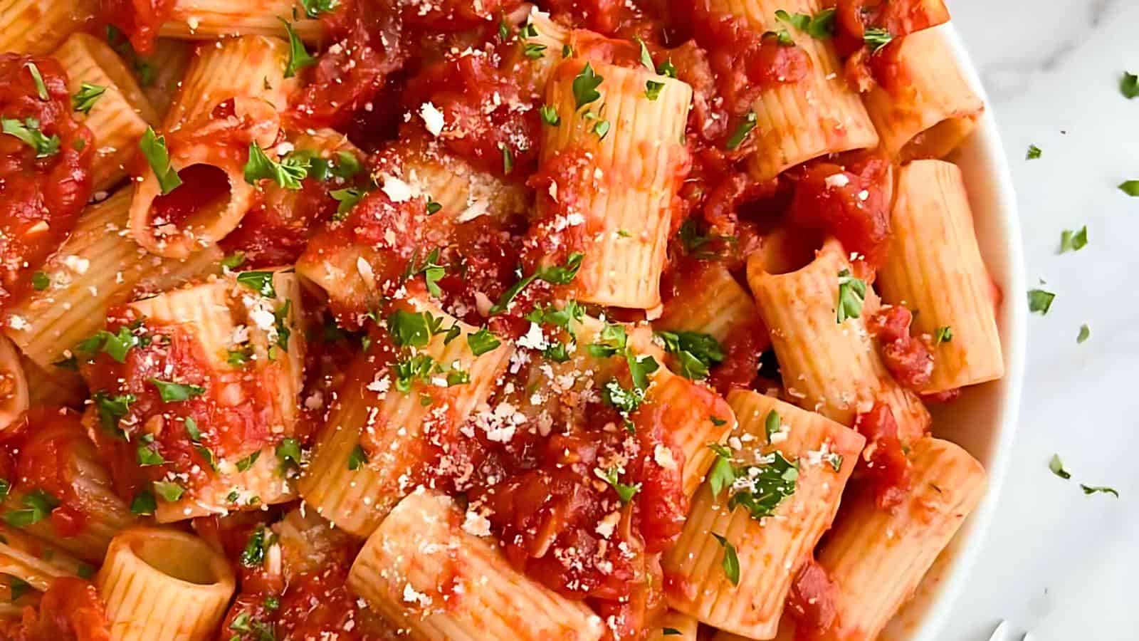 A close-up of a plate filled with rigatoni pasta covered in a chunky tomato sauce. The dish is garnished with chopped parsley and grated parmesan cheese, creating a colorful and appetizing presentation.